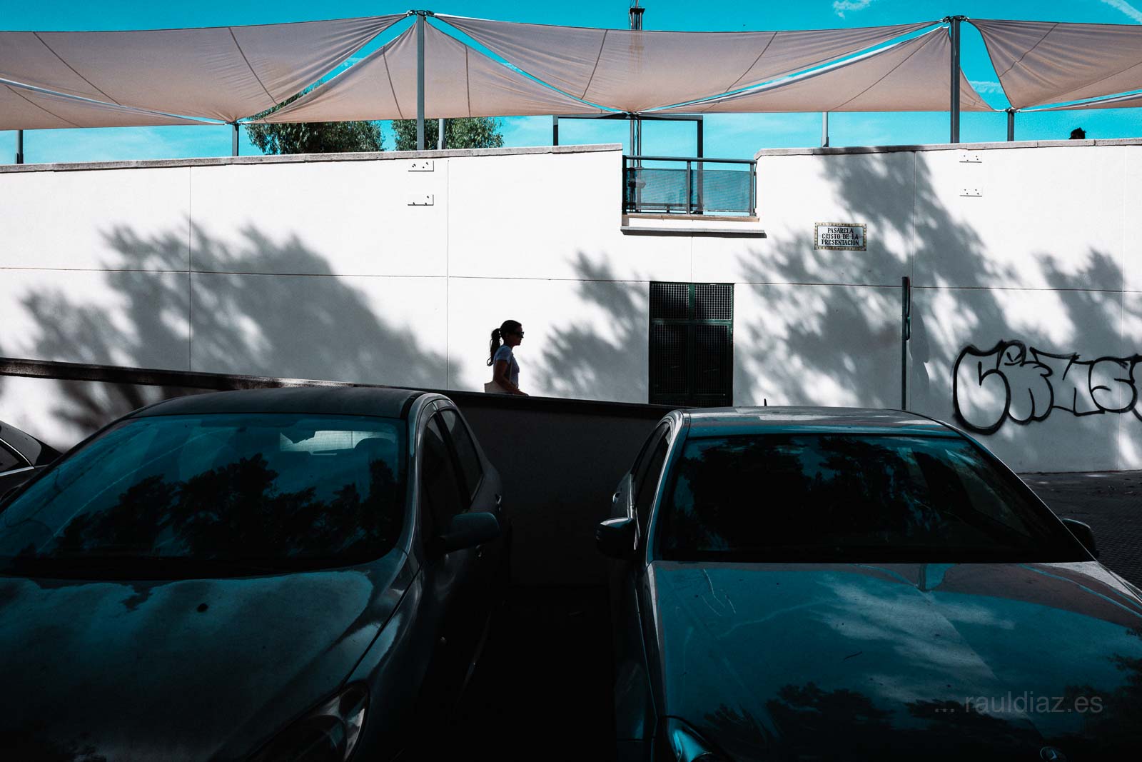 Mujer caminando por la Pasarela Cristo de la Presentación entre coches aparcados y sombras.