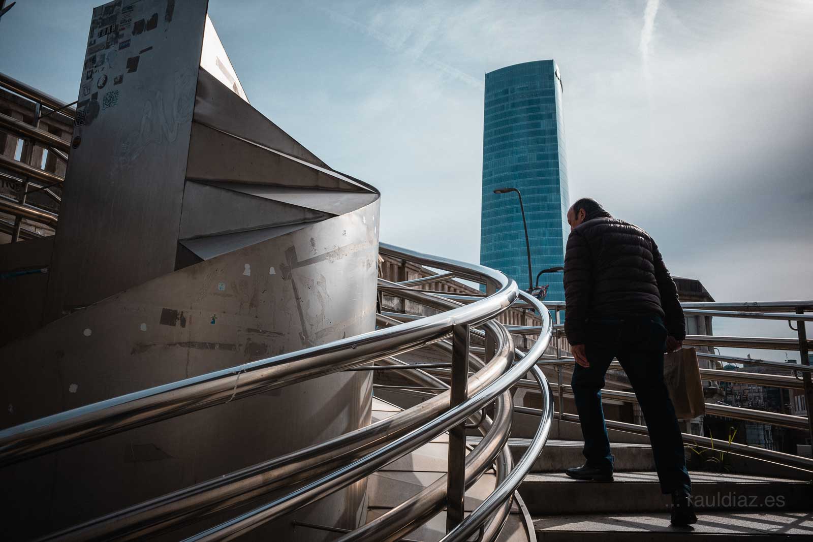 Hombre ascendiendo por la pasarela curva con la torre Isozaki al fondo