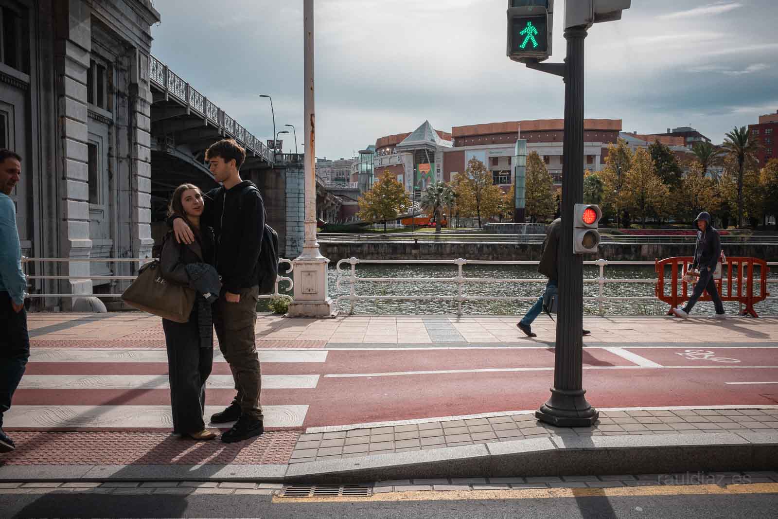 Pareja junto a la ría de Bilbao mientras otros peatones cruzan la escena