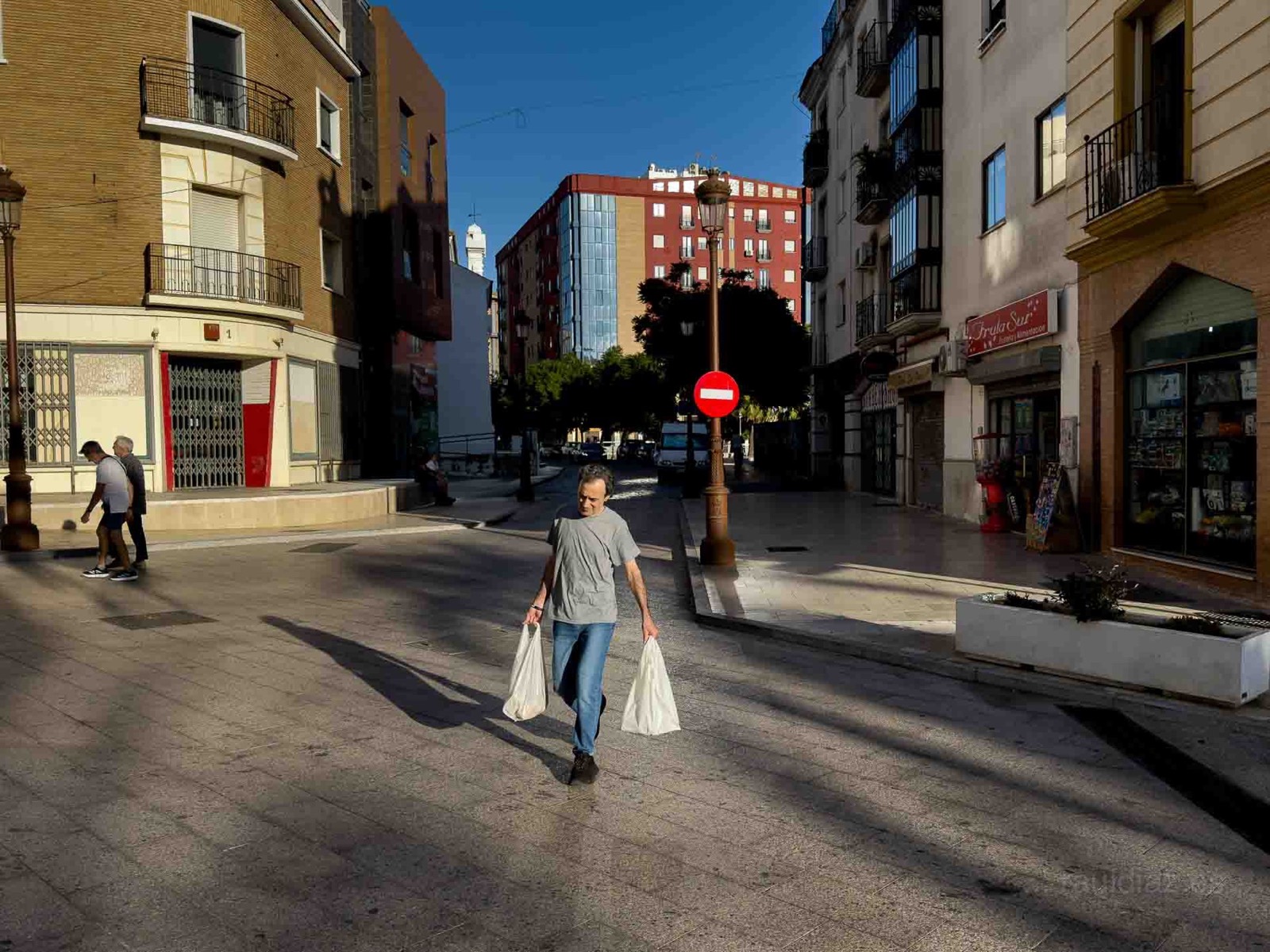 Hombre cruzando una calle con bolsas entre sombras, fotografía móvil en Huelva